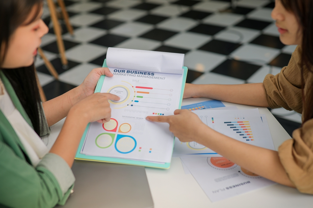 Two women talking to each other, pointing at different papers with colorful circles and bars. The title of a visible sheet says "our business, marketing management"
