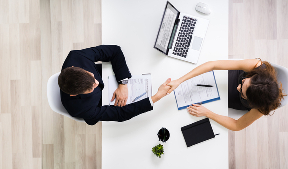Overhead view of a man and a woman shaking hands across the table. Printed sheets of paper, pens, and an open laptop on the table.