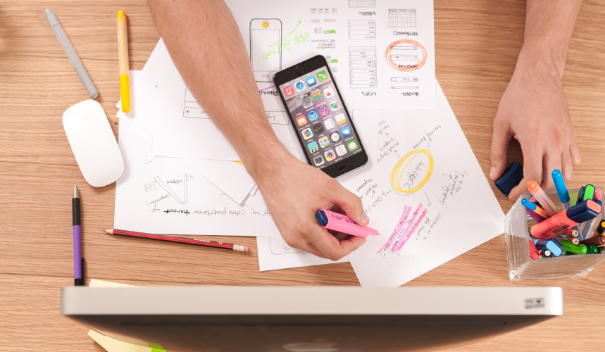 Overhead view of somebody working at a desk. Hands, paper, highlighters, cell phone and computer.